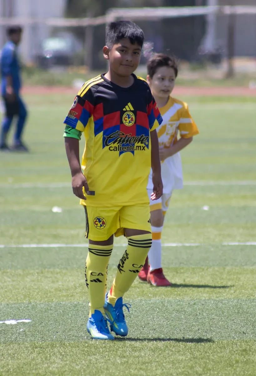 Niño de Nido Águila con uniforme amarillo, azul y rojo avanzando en el partido junto a un rival