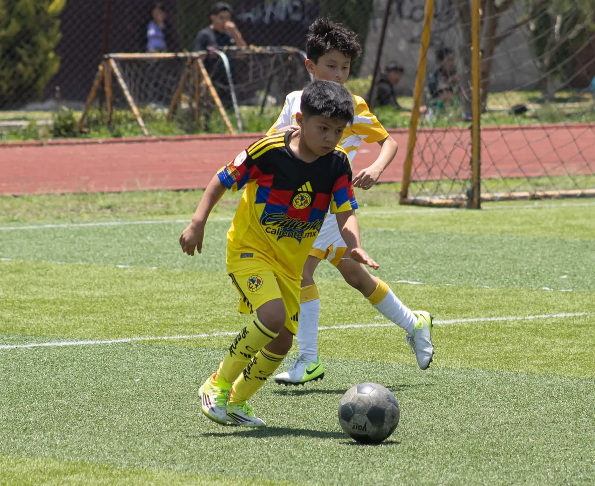 Niño de Nido Águila con uniforme amarillo, negro y rojo conduciendo el balón con un rival cerca
